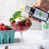 Person pouring olive oil over a dessert with strawberries and basil, with a bottle of Bona Furtuna olive oil in the background.