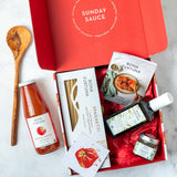 A curated Sunday Sauce gift box set from Bona Fortunea, featuring a bottle of marinara sauce, a box of spaghetti, a book, a small jar, and a bottle of olive oil arranged inside an open red box on a light marble surface next to a wooden spoon.