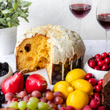 Panettone with white icing on a table with fruits and wine glasses.