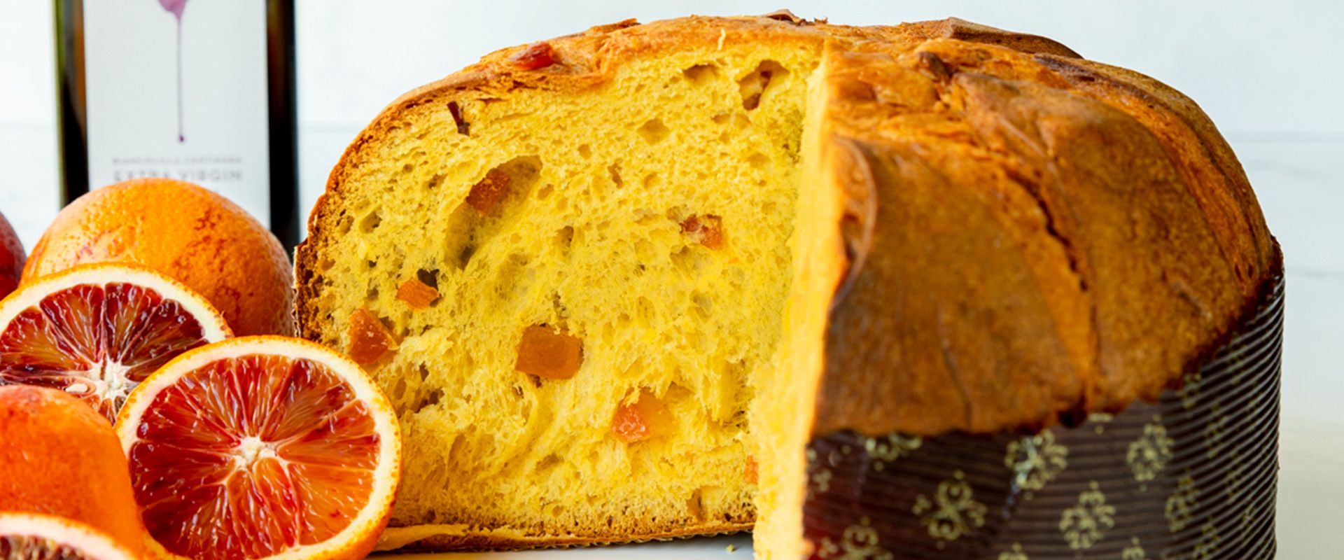 Loaf of panettone bread with a slice cut, surrounded by blood oranges on a white background