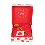 An open gift box containing a wooden spoon and a small card, with red tissue paper and a red inner lid. The lid is printed with the words 'SUNDAY SAUCE' in a circular design, and the outer box has a white base with red tomato illustrations.