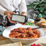 A person in a white shirt pours olive oil from a bottle labeled 'Pasta E Frittura' into a bowl of spaghetti with meatballs, set on a dining table with red wine glasses, bread, and a plant in the background.