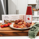 Bona Furtuna spaghetti pasta shown with a boxed package, a bottle of original pasta sauce, and a plated serving of spaghetti with meatballs on a wooden cutting board in a kitchen setting.