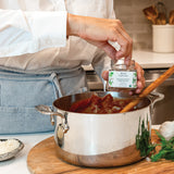 A person in a white chef's coat and blue apron adds a spoonful of pasta seasoning from a small glass jar labeled 'Pasta Perfuma' into a stainless steel pot of pasta on a wooden cutting board in a bright kitchen.